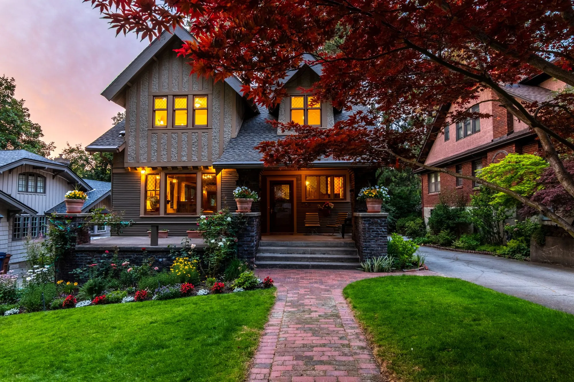 Craftsman house at dusk with lit windows, a stone porch and a brick path through a lawn