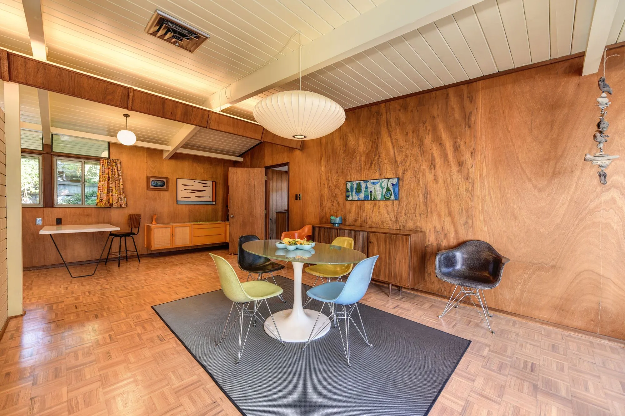 Wood-paneled dining room with a round table, colored chairs and a paper globe light